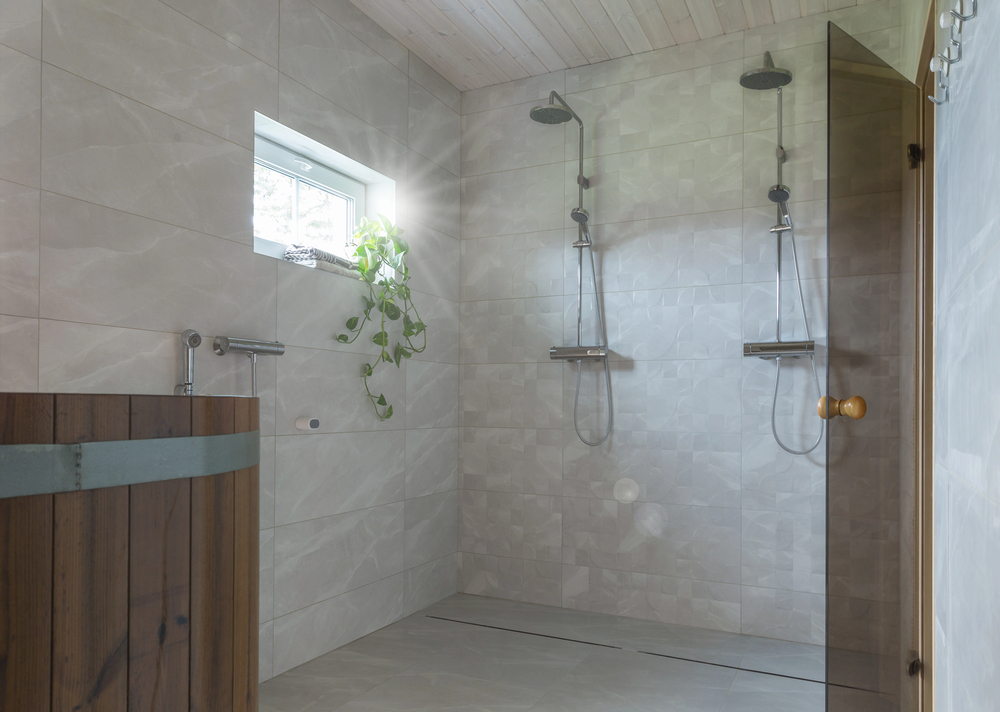 Modern bathroom by a leading construction company, featuring two wall-mounted showers, a small window with a leafy plant, tiled walls and floor, and a wooden tub in the corner.