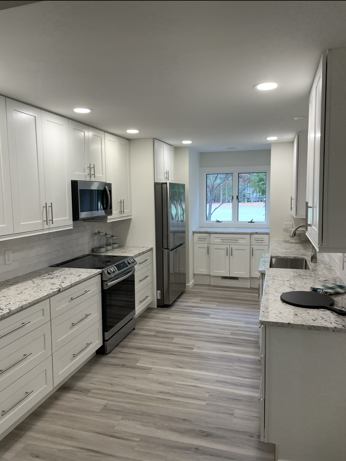 Modern kitchen with white cabinets, stainless steel appliances, light wood flooring, and large window overlooking a green outdoor area.