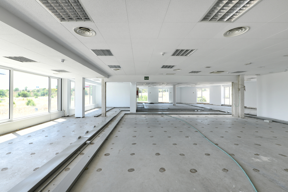 Empty office space with exposed floor panels, large windows, ceiling lights, and ventilation ducts, appearing to be under construction or renovation.