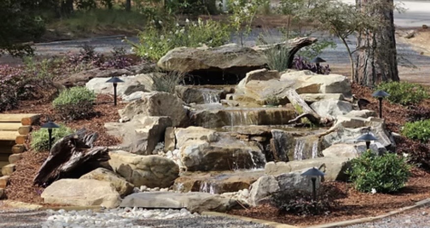 A small, tiered rock waterfall feature surrounded by mulch, shrubs, and landscape lights in a landscaped outdoor area.