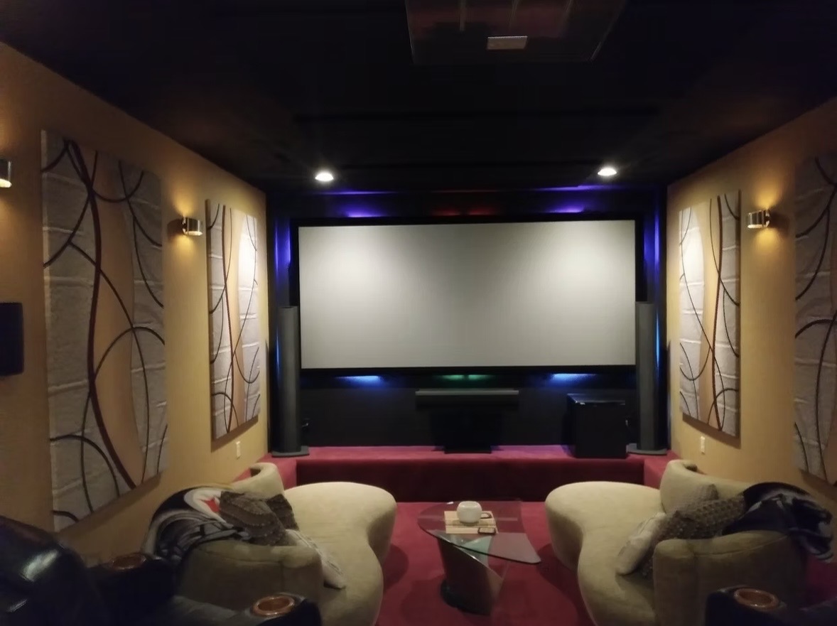 Home theater room with a large projection screen, two curved chairs, side tables, wall-mounted speakers, and decorative sound panels on tan walls.