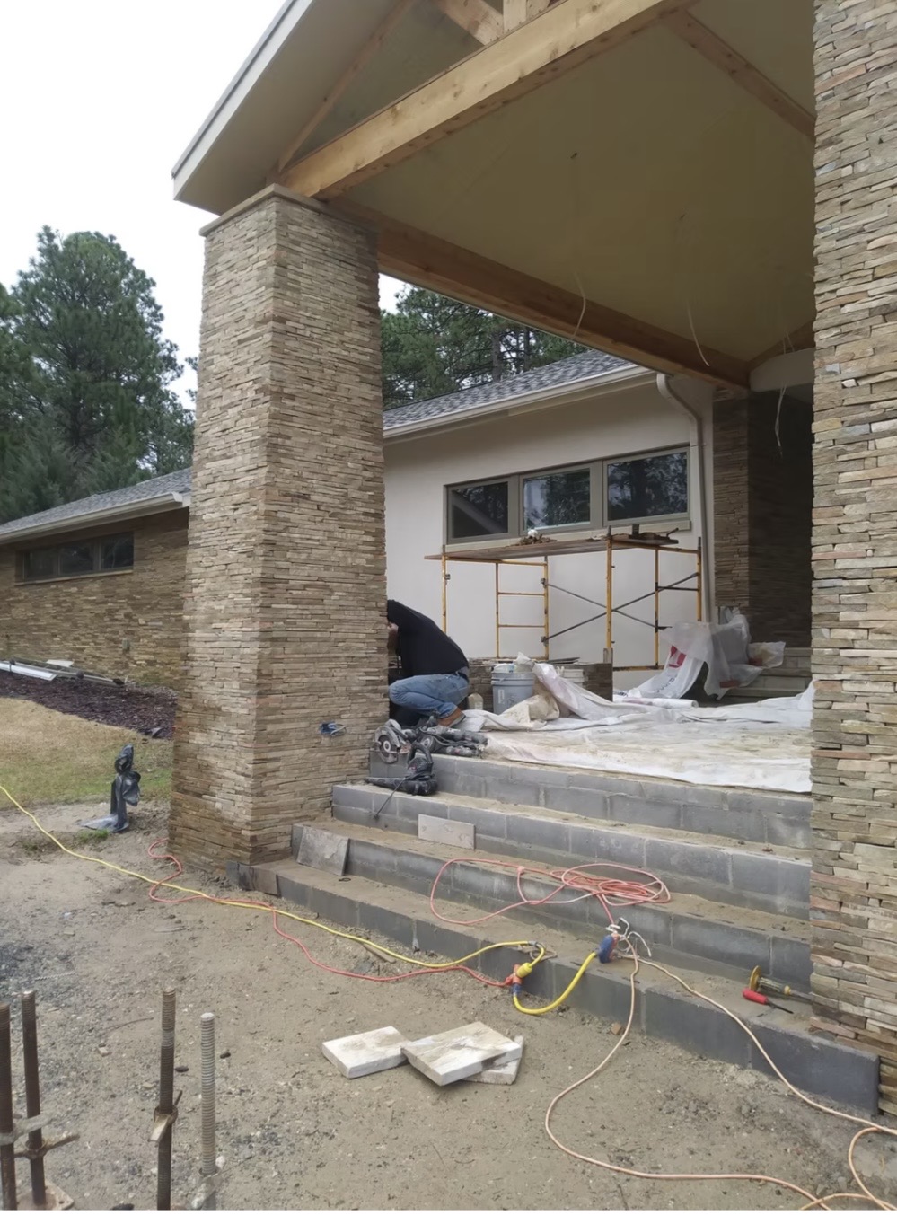 A person is working on the steps of a house under construction, with tools, cables, and scaffolding visible around the entrance.