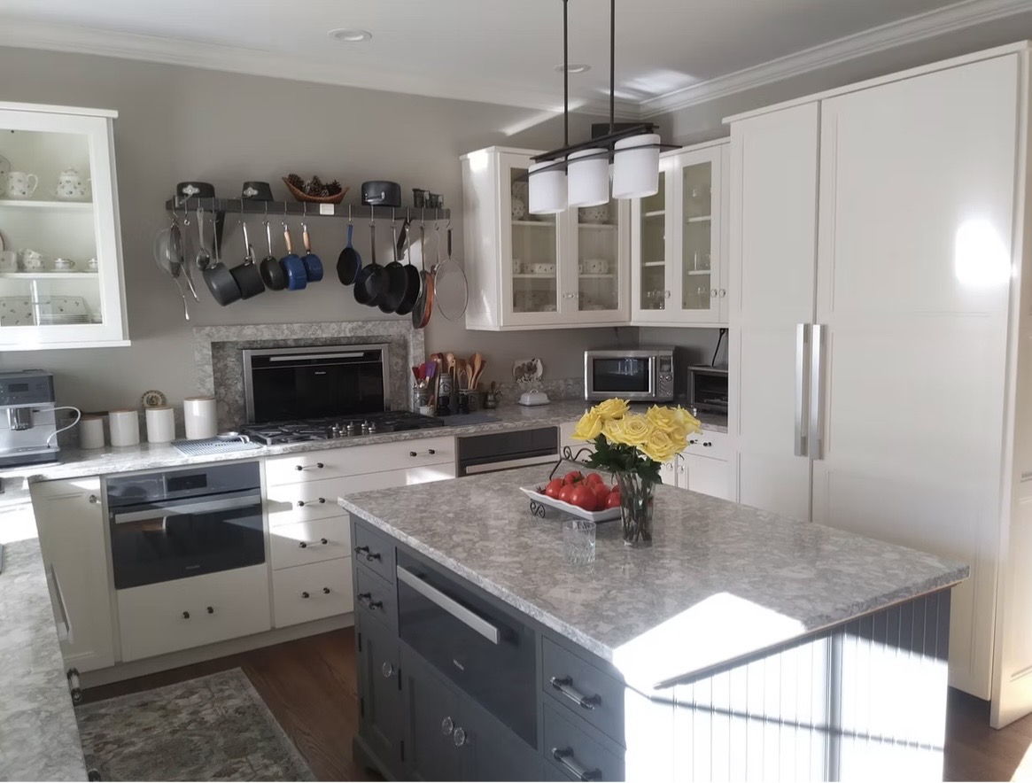 Modern kitchen with white cabinets, a marble island, stainless steel appliances, hanging pots and pans, and a vase of yellow roses on the island. Natural light fills the room.