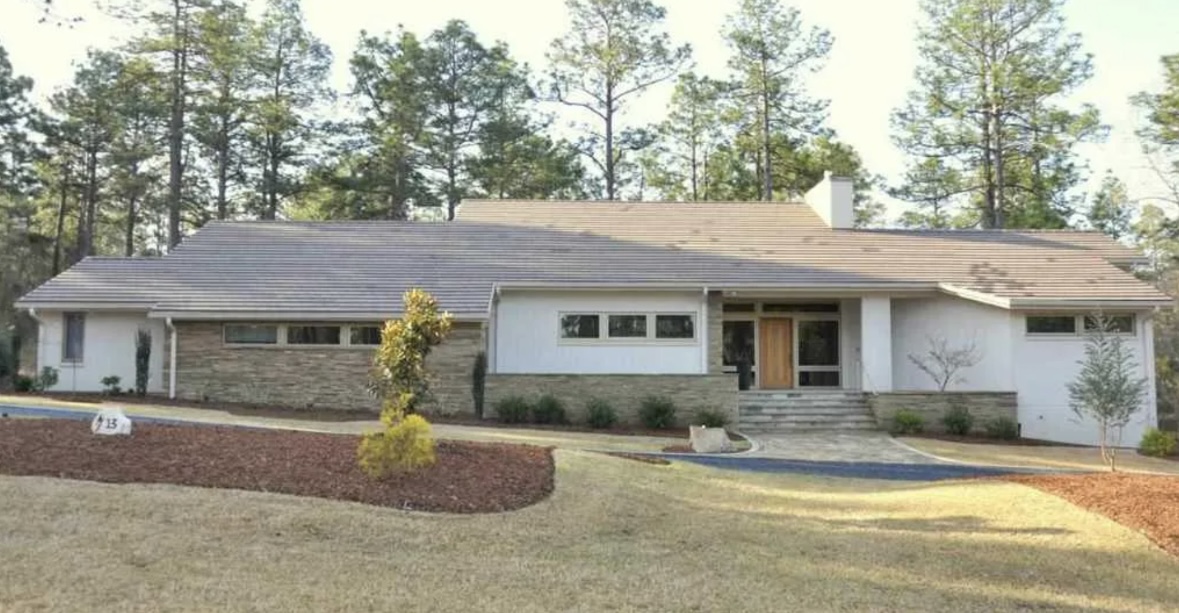 Single-story new construction home building with a sloped roof, stone and white exterior, front steps, and landscaped yard, surrounded by tall trees.