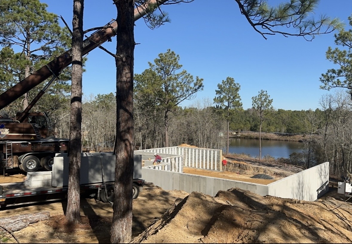 Construction workers build concrete walls for a foundation near a wooded area and a pond, with construction equipment and vehicles nearby.