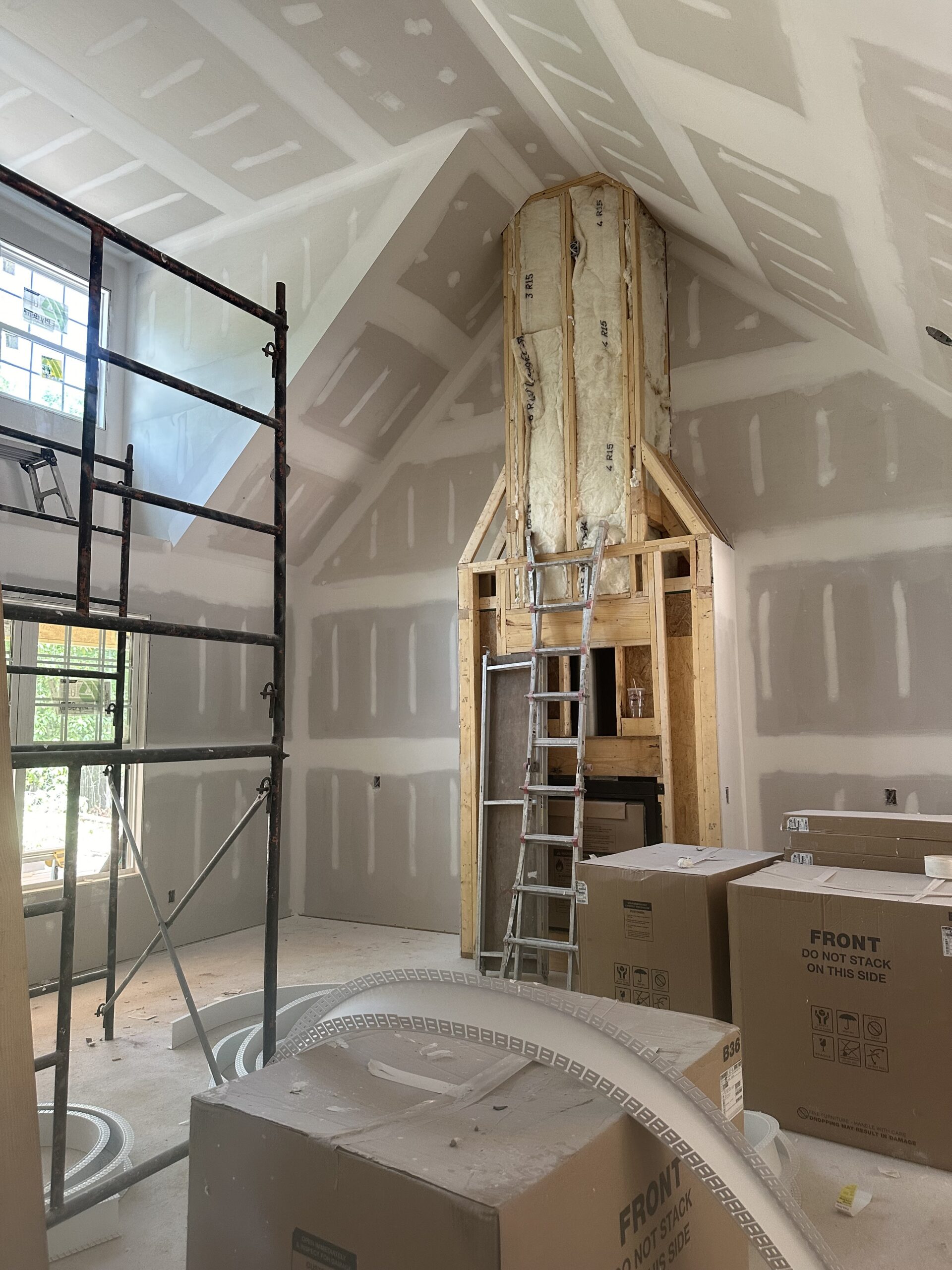 Interior of a house under construction with exposed drywall, insulation, a ladder, scaffolding, and several large cardboard boxes in the room.