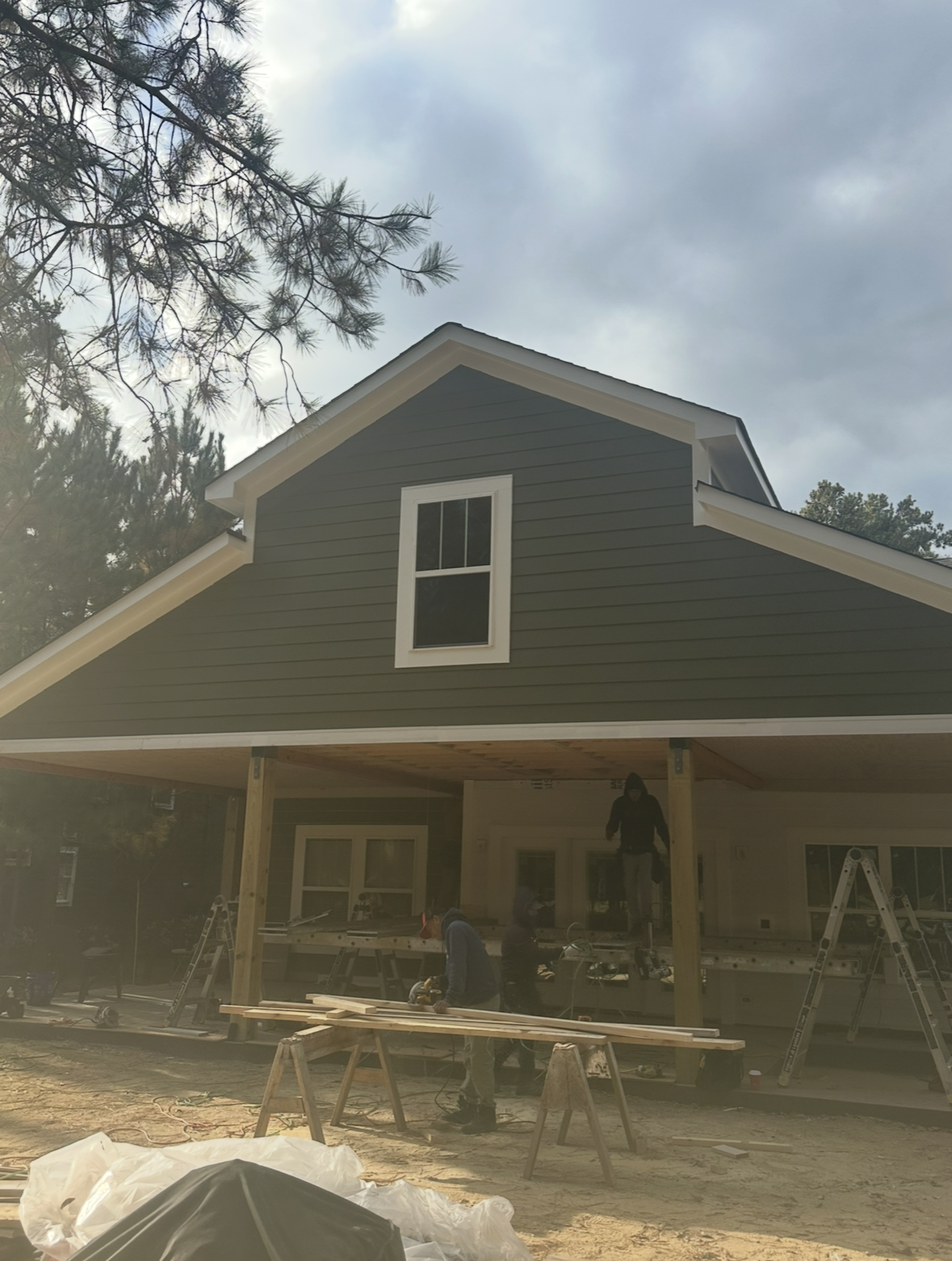 A green house with white trim under construction, featuring ladders and workers on the porch, with tools and materials scattered in the foreground.